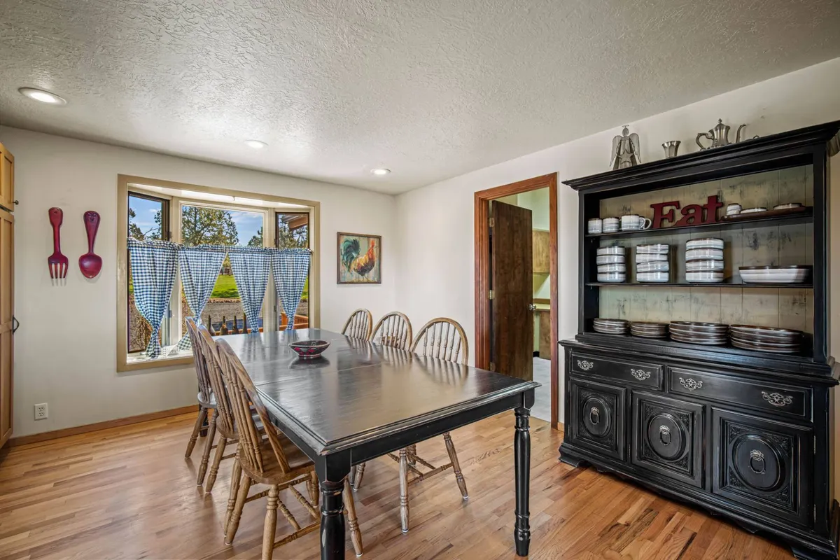 A cozy interior dining area with a central table, a black hutch, and a window dressed in checkered curtains. The setting is warm and inviting, ideal for family dinners or friendly gatherings. Board games or social activities could easily take place here, enhancing the communal aspect of the space.