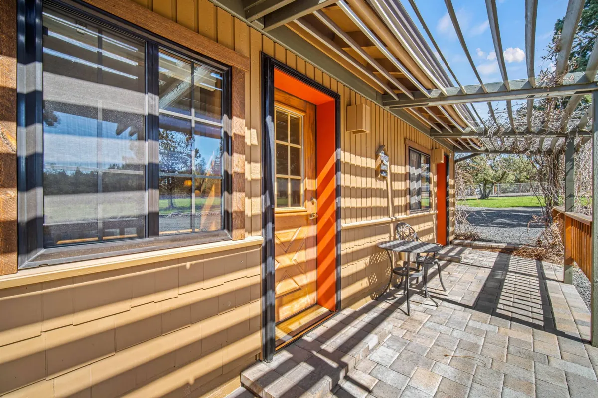 A bright orange door leads out to a shaded patio area with comfortable seating. The textured exterior façade offers a welcoming, contemporary vibe. The patio is surrounded by lush greenery, evoking a sense of relaxation and connection to the outdoors.