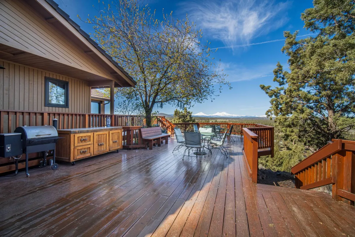 A wooden deck furnished with a dining set and a BBQ grill overlooks expansive mountain views and is framed by a stately tree. The setup clearly invites outdoor dining and relaxation, with nature all around and open sky above.