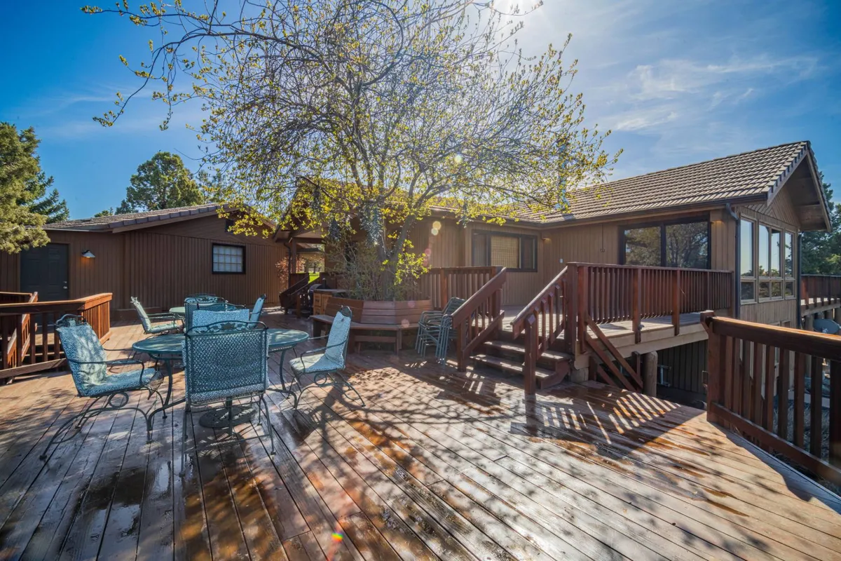 A tranquil exterior scene featuring a spacious deck beneath a large tree, arranged with an outdoor dining set. The deck is surrounded by greenery and offers plenty of room for group gatherings, meals, or simply relaxing in the sun. The distant view hints at scenic surroundings, ideal for sunset dinners or morning coffee.
