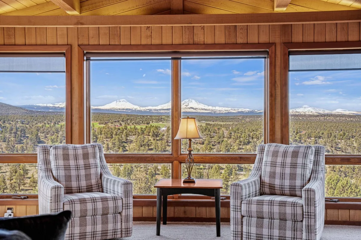 A sunlit seating area featuring large windows that frame sweeping mountain views. Plaid armchairs, a wooden accent table, and a soft lamp create a cozy, tranquil space perfect for morning coffee or conversation, while the room's open design connects directly to the outdoors.