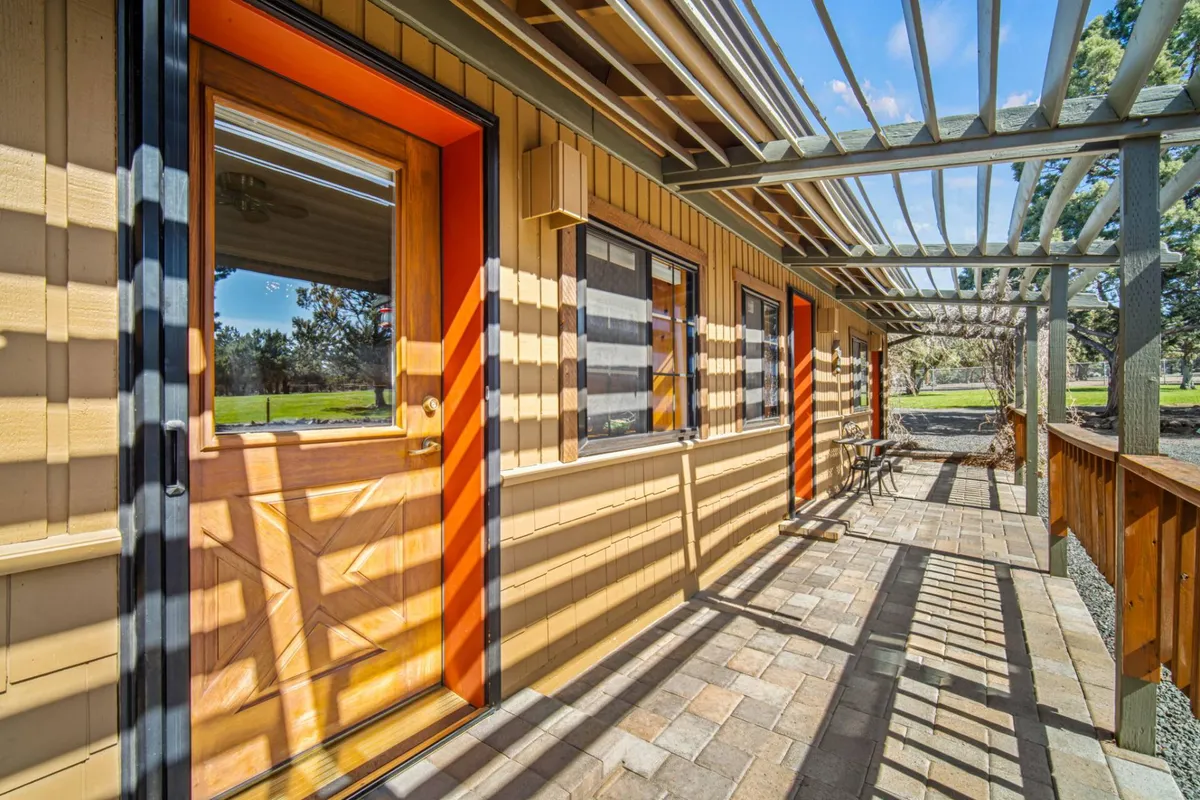 A paved pathway leads to an orange door beneath the shade of a pergola, with soft natural light and hints of garden views, suggesting a relaxing outdoor space connected to the home.