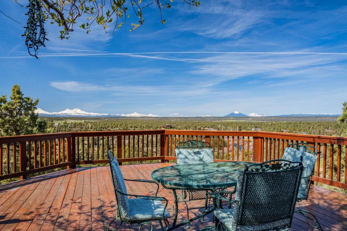 A deck equipped with a generously sized outdoor dining table, surrounded by a railing, sits against a stunning backdrop of a mountain range. The peaceful setting suggests both morning coffee moments and starlit evening gatherings.