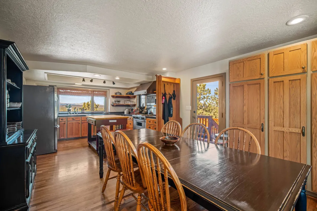 A spacious and inviting open-plan dining area and kitchen. The dining table sits beside a kitchen island, both set against a backdrop of wooden cabinets, stainless steel appliances, and a window offering scenic views. The ambiance is cozy and ideal for family meals or entertaining guests.