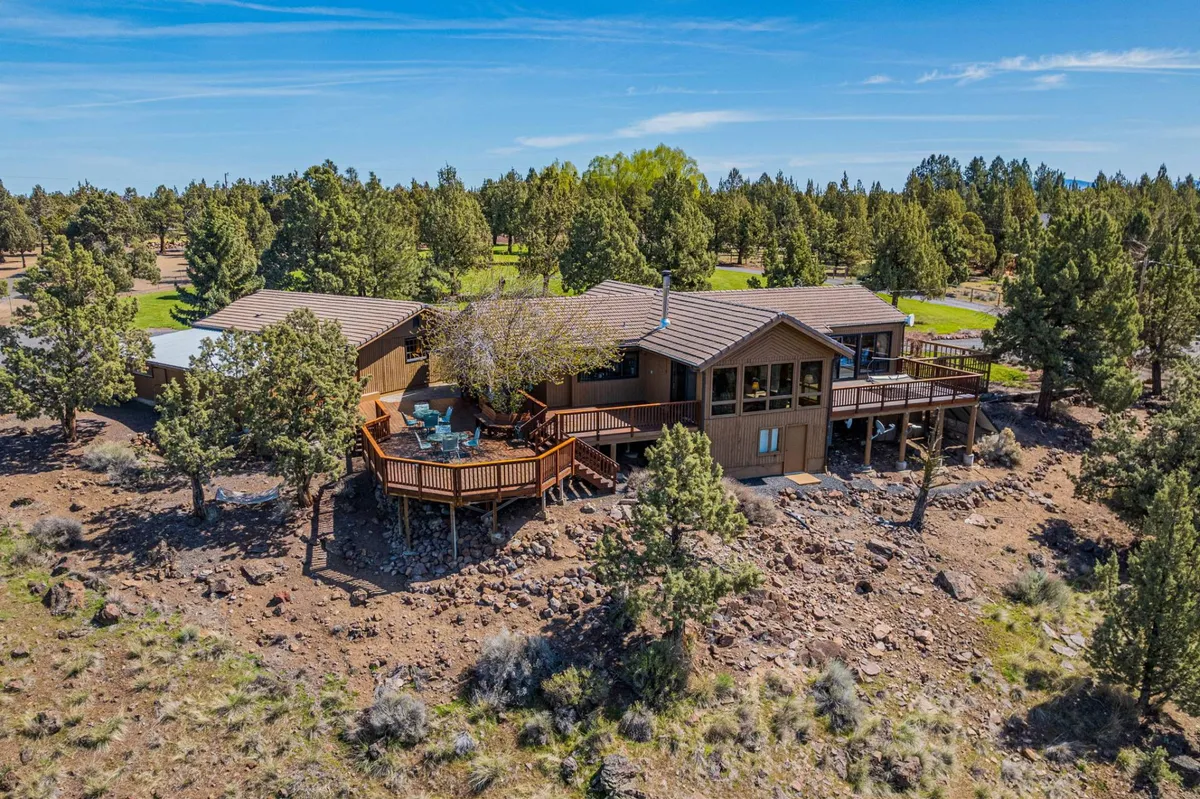 A spacious deck surrounded by trees and rocky landscape, with the main house structure visible. The deck offers plenty of room for outdoor gatherings, relaxation, and taking in natural surroundings. The setting is tranquil, with hints of sunset light and a sense of peaceful retreat.