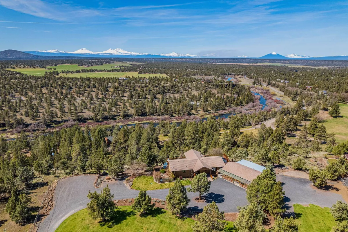 A sweeping view of the main house nestled beside a winding river, surrounded by lush forest and with distant mountains on the horizon. The landscape evokes the sense of being immersed in Central Oregon’s natural beauty, with clear skies overhead and a perfect mix of water, trees, and rugged high country.