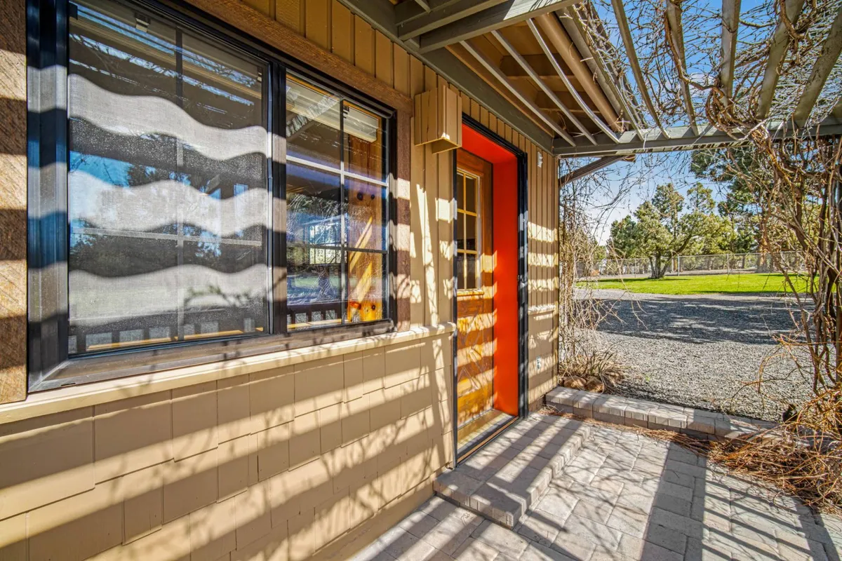 A vibrant orange front door stands out against a textured wall, with a window featuring striped curtains, suggesting a cheerful and welcoming entrance. The scene implies an entryway that blurs the line between indoors and out, inviting guests to step in or out as they begin or end their local adventures.