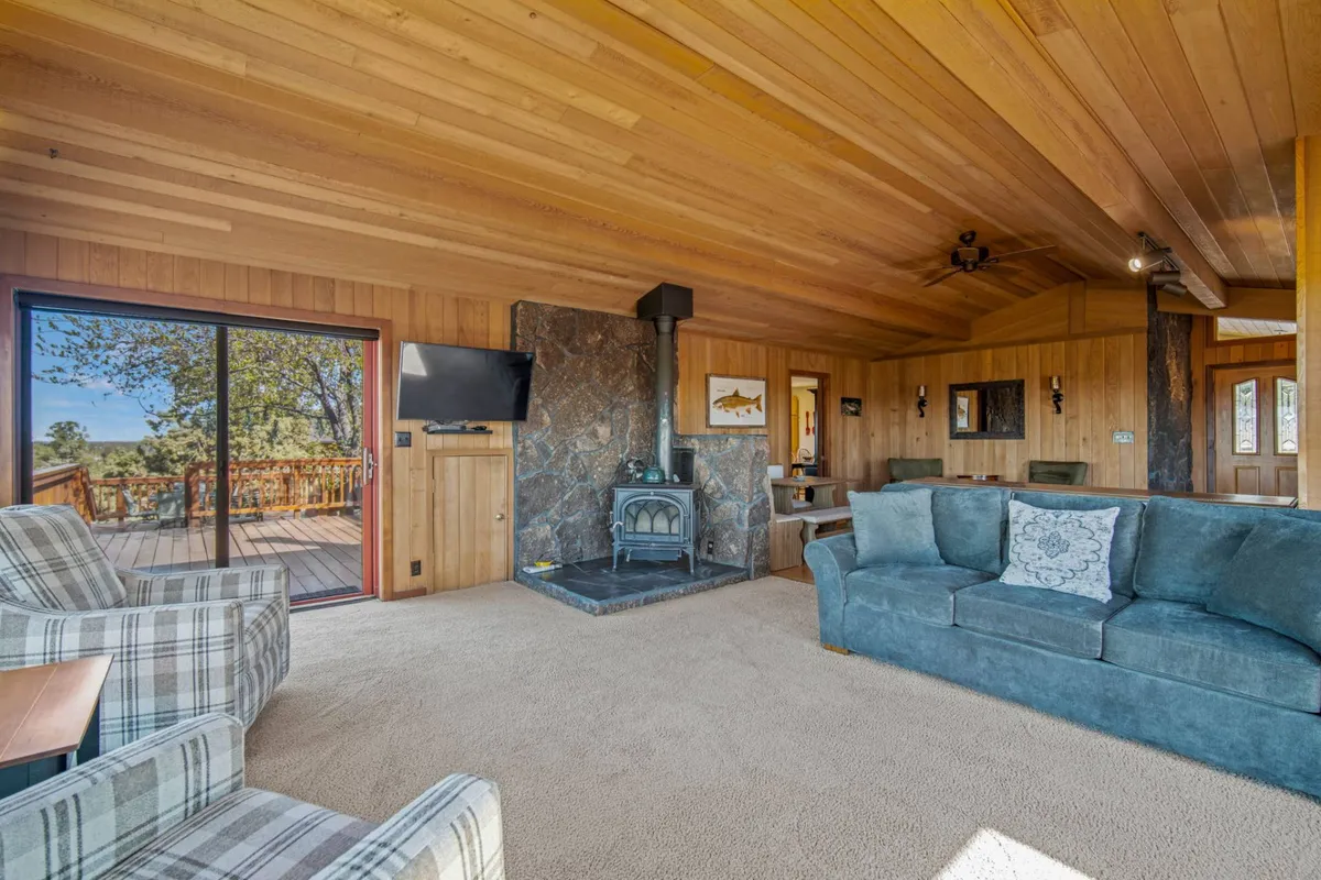 A welcoming living room anchored by a striking stone fireplace, wooden ceiling, plush blue couch, and plaid armchairs. Large windows bathe the space in light and offer glimpses of the landscape beyond. The room is designed for comfort, relaxation, and togetherness.
