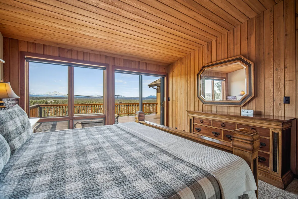 A serene bedroom with a comfortable bed situated next to large windows that frame breathtaking mountain views. The room is softly lit, with a dresser and a mirror creating a tranquil, restful atmosphere. Natural light pours in, highlighting the inviting simplicity and relaxation the space offers.