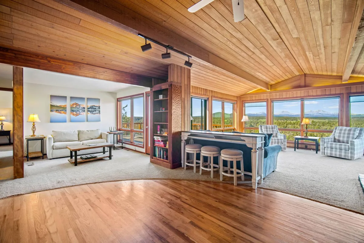 The image showcases a bright, spacious living area with soaring wooden ceilings, plush seating, and expansive windows framing breathtaking panoramic views of the Deschutes River and distant mountains. The cozy setup invites relaxation, family gatherings, and quiet moments spent soaking in the scenery or reading by the window, all bathed in natural light.